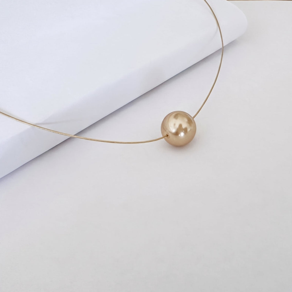 A delicate gold wire necklace with a single round, lustrous champagne-gold pearl pendant, displayed on a clean white surface.